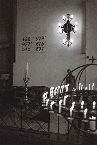 A grainy, black-and-white photograph from inside Oslo Cathedral. At the centre of the picture is the baptismal font from 1721. Above the font hang the numbers for the hymns of the day, and next to it stands a statue partially obscured by prayer candles in the foreground. The mood is solemn, as attested by the burning candles and the statue's slightly lowered head.