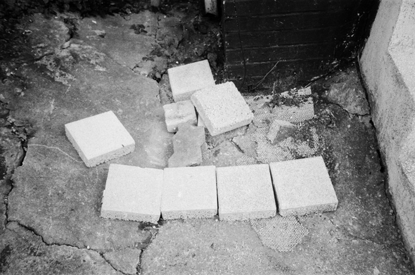 깨지고 낡은 콘크리트 위에 정사각형 모양의 콘크리트 타일 4개가 나란히 놓여있다. 그 왼쪽 위에 삐뚤게 타일 하나가, 그 위쪽에 타일 두어 개가 놓여있다.