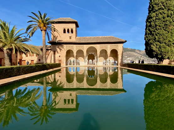 One of the buildings of Alhambra, palm tree on the left, pine tree on the right, clear blue sky, all perfectly mirrored in the small pond before it.