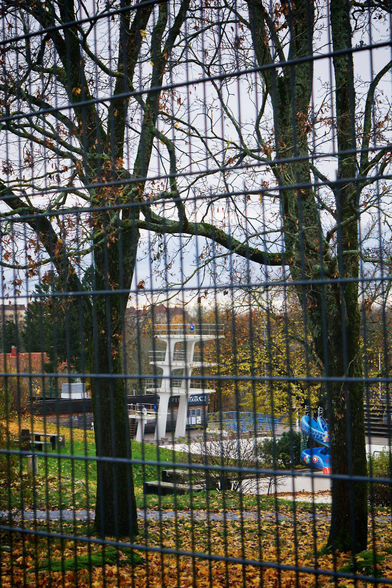 The diving tower of the outdoor swimming pool stands alone at the edge of the empty pool on the Samppalinna hill in Turku, Finland. Autumn leaves lie scattered here and there. The photo was taken from behind a safety fence, its mesh blurred in the foreground. Two mostly leafless deciduous trees by the fence frame the diving tower.