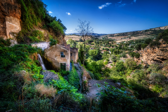 Farbfotografie einer verlassenen oder baufälligen Gebäudegruppe, eingebettet in eine steile, felsige und üppig bewachsene Landschaft, wahrscheinlich in einer Schlucht oder einem Tal.

Die Gebäude, deren Fassaden hellbraun bis ockerfarben sind und teilweise zerfallen, schmiegen sich links an eine hohe, rötliche Felswand. Links neben dem vorderen Haus fliesst ein kleiner Wasserfall über bemooste Steine hinab.

Die gesamte Umgebung ist von dichtem, dunkelgrünem Gebüsch und Bäumen überwuchert, was der Szene einen verwunschenen Eindruck verleiht. Ein einzelner, kahler Baumstamm ohne Blätter steht in der Mitte zwischen den Ruinen und dem Hintergrund.

Im Hintergrund öffnet sich die Sicht auf eine weite, hügelige Landschaft mit vereinzelter Vegetation, Feldern und weiteren Felsformationen. Der Himmel ist tiefblau mit einigen weissen Wolken.