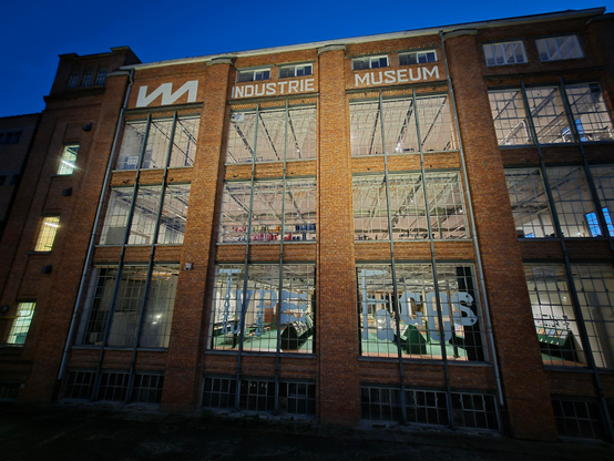 Hoge, oranje, bakstenen façade met wit opschrift "Industriemuseum". Foto genomen in vooravond, met een diepblauwe hemel zichtbaar achter het gebouw. Door de grote raampartijen zijn de verlichte expositieruimten zichtbaar.