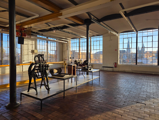 Open ruimte in een industrieel pand (Industriemuseum Gent), waarbij het zonlicht binnenvalt door de grote ramen en lange schaduwen werpt op de bruin-oranje tegelvloer. Centraal-links zijn enkele zwart-metalen voorwerpen zichtbaar. Achteraan een wit geschilderde bakstenen muur met gele accenten. Door de ramen zie je de blauwe hemel en de bekende torens van Gent.