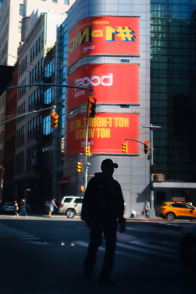 A silhouette is seen through the reflection of a window. Behind them towers three large orange billboards on the side of a gray metal building.