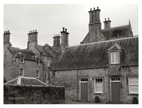 Black and white photo of historic stone buildings with multiple tall chimneys and steep roofs. Closed shutters and small doors convey an old-world charm.