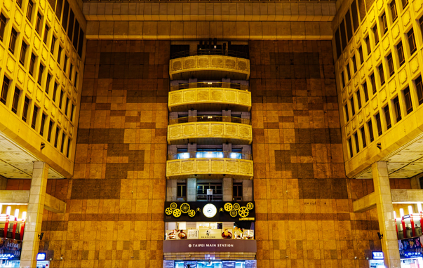Wall with balconies, a clock, clockwork decoration, and a sign that reads “Taipei Main Station” inside an atrium-like ticketing hall.