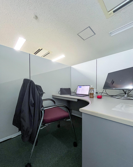 Mi cubículo dentro de la sala común. Una silla (con la chaqueta en la parte de atrás) y una mesa rodeada de paredes de plástico gris con una altura de un metro y poco. Sobre la mesa una tablet, un portátil (encendido), una botella de café con leche (Boss), la funda de las gafas y una pantalla grande (que no llegué a usar).
