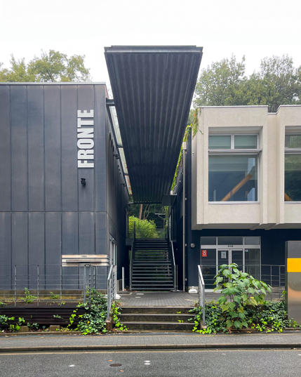 Un edificio con un diseño llamativo dentro del campus. Se ve una escalera que parece llevar a un bosque con una cubierta estrecha y alargada. A los lados hay un edificio con ventanales (¿una biblioteca?) y otro sin ventanas en cuya fachada pone FRONTE (no sé qué era).