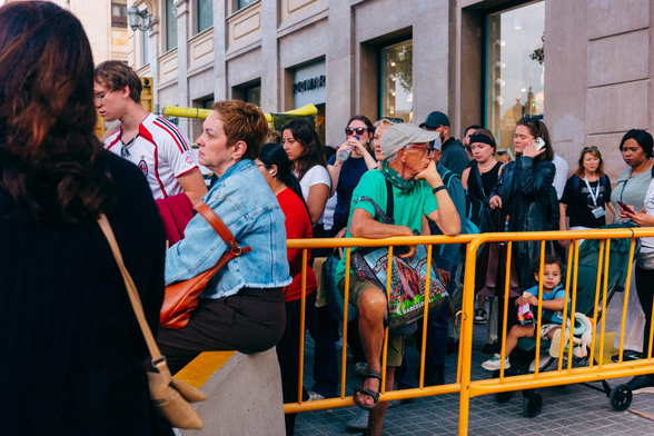 Fotografía de una calle del centro de Barcelona con vallas, gente sentada o apoyada en ellas, gente tratando de pasar, y todo el mundo con cara de no estar pasándoselo nada bien.

Photograph of a street in the center of Barcelona with fences, people sitting or leaning on them, people trying to get through, and everyone looking like they're not having a good time at all.
