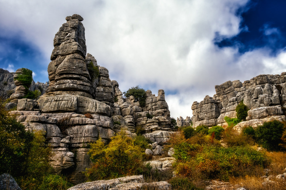 Farbfotografie einer surreal wirkenden Karstlandschaft, dominiert von bizarren, gestapelten Felsformationen.

Die Szene wird von hohen, säulenartigen Kalksteintürmen beherrscht, die durch horizontale Verwitterungsschichten zerfurcht sind. Der grösste Felsen im linken Vordergrund ragt massiv in die Höhe und ist an einigen Stellen leicht von Pflanzen bewachsen.

Die Felsen haben eine hellgraue bis beige Farbe. Im Vordergrund und im unteren Teil des Bildes gibt es dichte, buschige Vegetation in Herbstfarben: Grüntöne mischen sich mit leuchtendem Gelb und Rostrot.

Der Himmel ist dramatisch, mit tiefblauem Himmel, der von grossen, bauschigen weissen und grauen Wolken durchzogen wird. Das Licht ist kontrastreich, was die Textur der Felsen stark betont und dem Bild eine düstere, gleichzeitig majestätische Stimmung verleiht.
