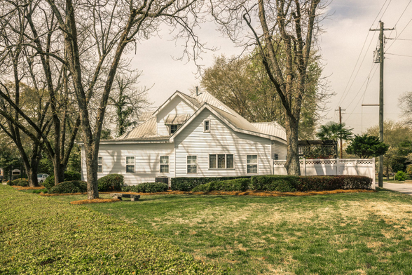 White clapboard farmhouse with pale gray metal roofing and multiple steep gables. The two-story home features symmetrical windows with white trim and sits on a spacious lawn bordered by a low hedge of rounded shrubs. Mature bare deciduous trees frame both sides of the property, with moss-covered trunks visible in the foreground. A white decorative fence with lattice details is visible on the right side of the property. Utility poles and power lines cross the overcast sky in the background. The lawn shows patches of early spring growth with shadows cast across the grass from the surrounding trees.