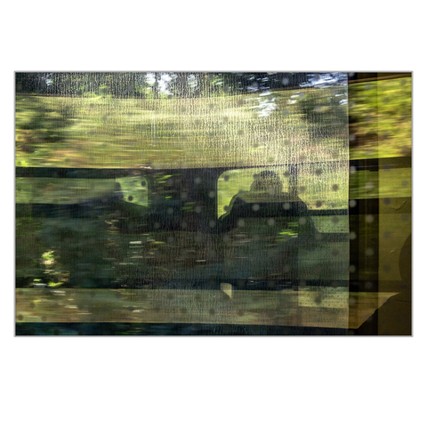 The speckled window of a train compartment reflects the window of the train corridor, through which a man looks out, with passing, blurred trees visible in the background.