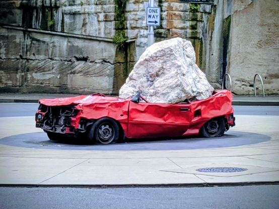 Ah, Still Life with Stone and Car by Jimmie Durham, a provocative and deeply resonant piece. Here, Durham challenges the very foundations of Western sculptural tradition, eschewing the heroic bronze and marble for the abject detritus of our consumer-driven society. The automobile, that quintessential symbol of progress and personal freedom, is rendered a crushed, impotent husk, its vibrant red a jarring counterpoint to its brutalized form.

And then, the stone. Not a chiseled, idealized form, but a raw, unyielding mass, asserting its primal presence with an almost brutalist honesty. It rests upon the car not merely as an object, but as a silent, geologic force, suggesting a profound reordering of power dynamics. Is this nature reclaiming its dominion over humanity's manufactured hubris? Or perhaps a wry commentary on the weight of history and expectation crushing the contemporary?

Durham, with his characteristic wit and anthropological insight, forces us to confront our own material desires and the often-overlooked violence inherent in our built environment. The street art context further amplifies its message, transforming a public thoroughfare into a transient gallery, democratizing the act of critical engagement. A truly masterful juxtaposition of the organic and the manufactured, the monumental and the mundane, compelling us to re-evaluate our perceived realities.

AltText created by Gemini Flash 2.5 as a pretentious art critic and checked with a smile by human.