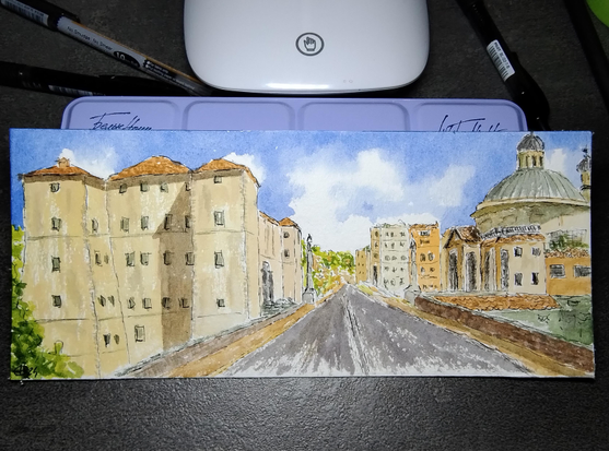 Italiano:
Un dipinto acquerello mostra una strada cittadina fiancheggiata da alti edifici color giallo e beige. Sulla destra, una grande chiesa con cupola verde spicca tra le case. Il cielo è azzurro, punteggiato da nuvole bianche. La prospettiva porta lo sguardo verso il fondo strada, dove la vegetazione appare tra altri palazzi. I dettagli sono delicati, gli edifici presentano numerose finestre.

English:
A watercolor painting depicts a city street lined with tall yellow and beige buildings. On the right, a large church with a green dome stands out among the houses. The sky is blue with white clouds. The perspective leads the eye down the road, where greenery appears between more buildings. Details are delicate, with many windows on the buildings.