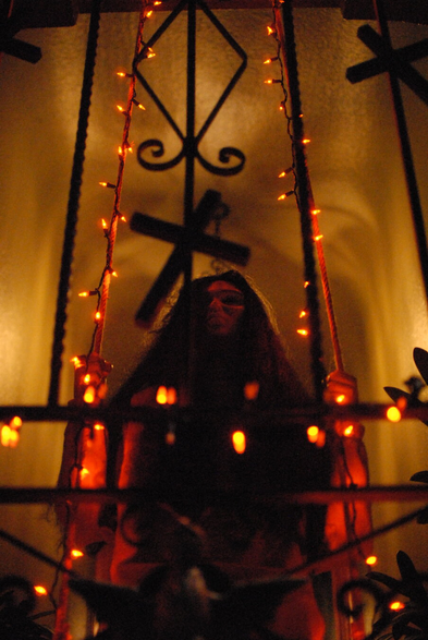 A mysterious woman sits on a swing behind a gate. She's backlit by a dim light.