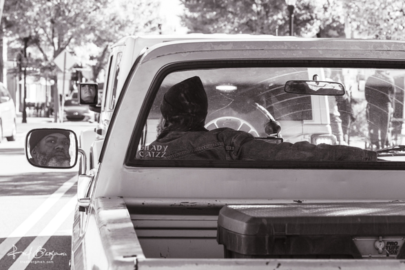 Man in pickup truck photographed from behind while his face is reflected in the side view mirror and the rear window has sticker that says "Shady Catzz"