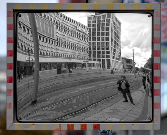 Foto eines Spiegels für die Straßenbahnfahrerin an einer Haltestelle. Der Spiegel hat eine verblasste rot-weiße Umrandung. Im Spiegel sieht man den Fotografen, die Gleise und zwei Gebäude im Stil der 70. Jahre
