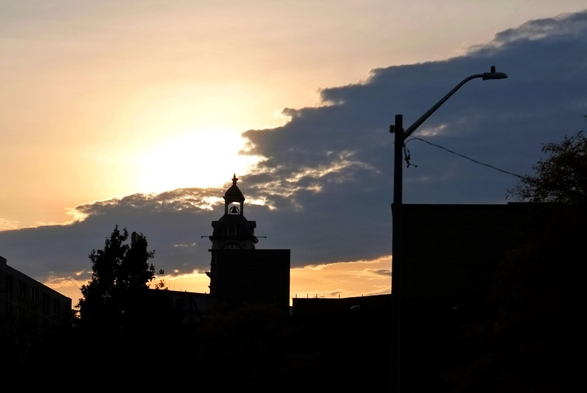 The setting sun is seen partially hidden by a large cloud in a golden yellow sky. The silhouette of buildings, trees and a light pole is seen along the bottom.