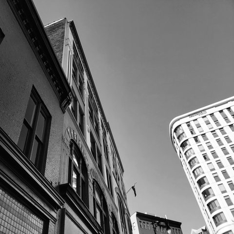 Low-angle, black and white photograph of historic brick buildings under a clear sky. The left side showcases a detailed facade with large windows and ornate trim, while the right features a curved, modern high-rise building. A flag waves in the breeze.