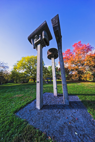 Three concrete stems with square and round things on top rise out of a gravel patch in the middle of a green lawn with strident red and green trees in the background under a clear blue sky