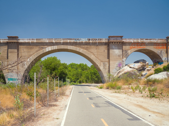 A concrete bike path and a dry river pass under the great arch of a bridge