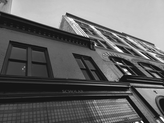 Low-angle shot of an old brick building with "SCHOLAR" displayed on a lower facade, featuring dark windows and ornate detailing, with the building's facade angled toward the sky.