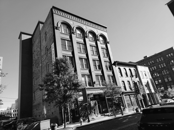 Black and white street-level photograph of a multi-story brick building with arched windows on the upper floors. A brick facade runs down the side of the building with the word "SEARS" printed vertically. Adjacent buildings are visible, as well as a street with a car and trees. The perspective is tilted upward.