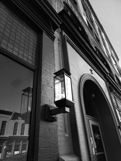 Low-angle, black and white shot of an old brick building exterior featuring a vintage lantern mounted on the wall. The lantern, enclosed in a rectangular glass case, casts a bright light. Beside the lantern is a large window with a pane of glass that reflects another building across the street. Further down the building, an arched doorway leads to the Grand Rapids Brewing Co., and a glimpse of the sky is seen.