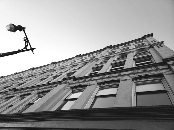 Low-angle black and white photograph of a tall brick building with many rectangular windows, perspective converging upwards, with a decorative wrought-iron street lamp in the left foreground and a clear sky in the background.