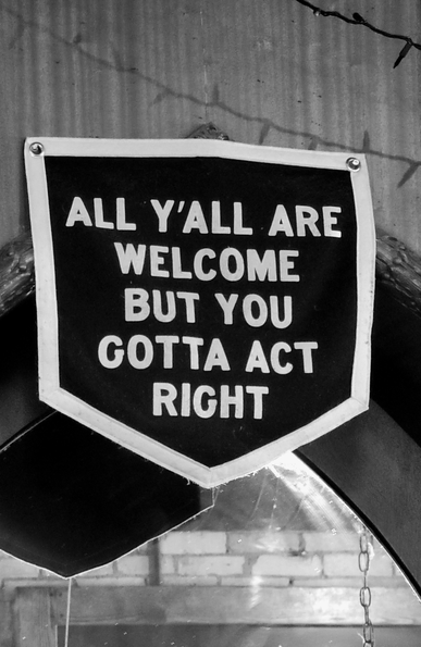 Black and white photograph of a sign with a white border and the words "ALL Y'ALL ARE WELCOME BUT YOU GOTTA ACT RIGHT" in white text on a black background, with a reflective surface and a brick wall visible beneath.
