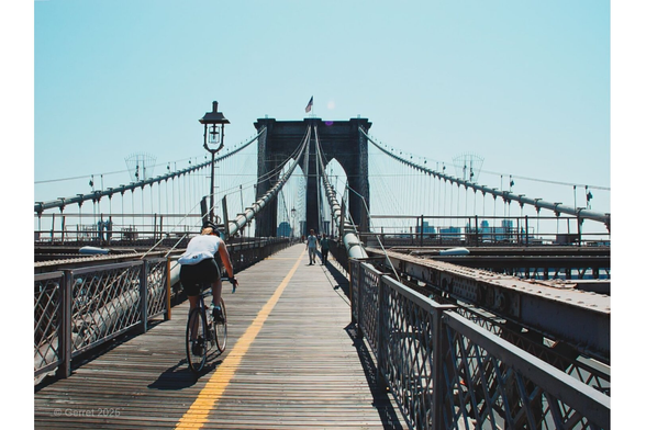 Cyclist riding on a wooden pedestrian path of the Brooklyn Bridge, under a clear blue sky, with the iconic suspension cables and distant city skyline.