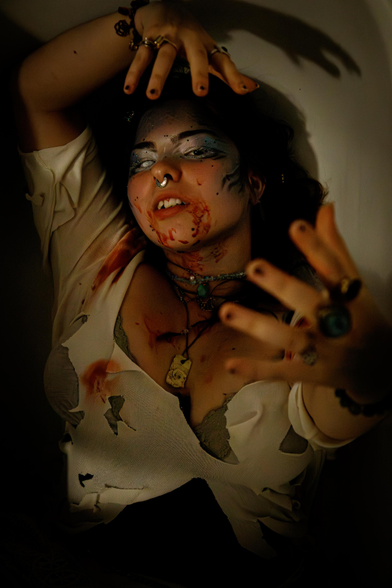 A siren lies in dim light with torn clothes and dramatic makeup, including fake blood on her face and chest. She wears jewelry and reaches towards the camera, creating a dark, eerie atmosphere.