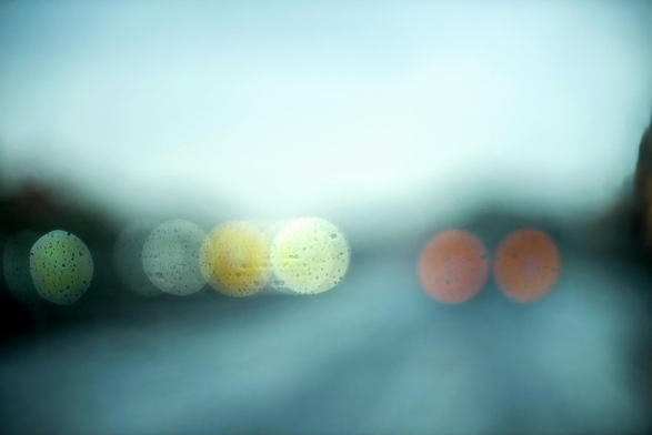 Das Bild zeigt eine Aufnahme durch die Frontscheibe eines Fahrzeug bei Regen.  Die Scheinwerfer und Rücklichter der anderen Fahrzeuge, sind als runde Kreise mit Wassertropfen darin zu erkennen.