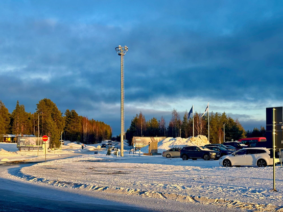 Clear sunset light on a snow covered parking lot, a forest of tall spruces cut by a road in the foreground, cloudy sky with a few openings of blue.