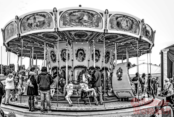 Schwarzweißfotografie im Format 3:2 Landscape.
Ein original restauriertes, altes Kinderkarussell steht auf einem Volksfest. Pferde und Kutschen sind aufwendig und liebevoll gestaltet. Das Dach und die Wände des inneren Karussellkreises sind ebenfalls kunstvoll und handbemalt.