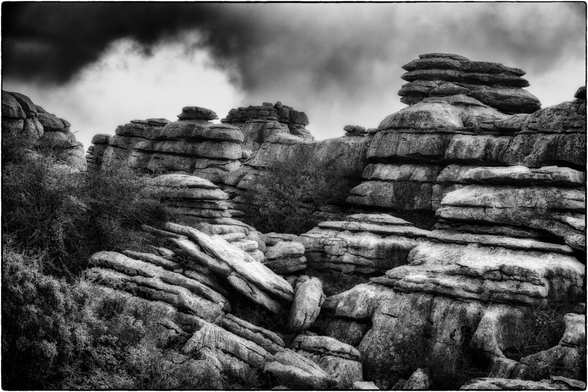 Schwarz-Weiss-Aufnahme einer wilden Karstlandschaft, dominiert von horizontal geschichteten Felsformationen unter einem dramatischen Himmel.

Im Vordergrund erstreckt sich eine Masse aus grossen, flachen und zerbrochenen Kalksteinplatten, die schräg in die Tiefe führen. Die Textur der Felsen ist rau und detailliert, mit dunklen Rissen und hellen Oberflächen.

Im Mittel- und Hintergrund türmen sich gestapelte Felsformationen auf, die wie natürliche Steinmauern oder Säulen aussehen, typisch für eine Karsthöhenlandschaft. Die Linien der Felsenschichten sind klar erkennbar.

Zwischen den Steinen wächst spärliches, struppiges Gebüsch. Der Himmel ist sehr kontrastreich, mit dunklen, dramatisch gezeichneten Gewitterwolken, die die gesamte Szene bedrohlich wirken lassen. Die Vignettierung am Bildrand verstärkt diesen düsteren Eindruck.