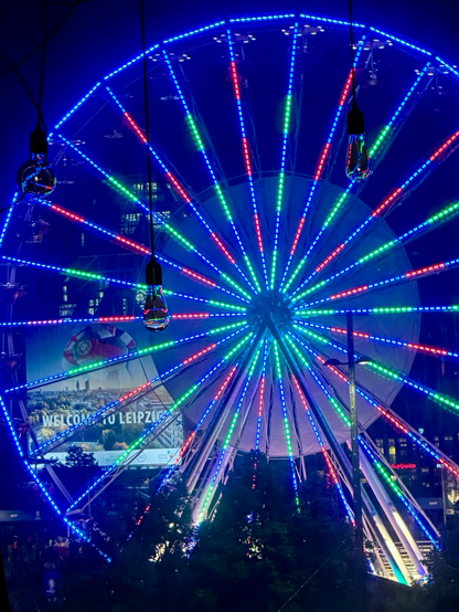 Ein beleuchtetes Riesenrad in der Nacht. Das Rad deckt das ganze Bild ab. Es leuchtet blau, rot, grün. Im Vordergrund hängen drei Glühbirnen, da das Foto durch das Hotelfenster gemacht wurde. 