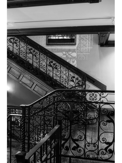 Black and white photo of an ornate, wrought iron staircase with intricate scrollwork. Soft light casts shadows on the walls, creating a nostalgic atmosphere.
