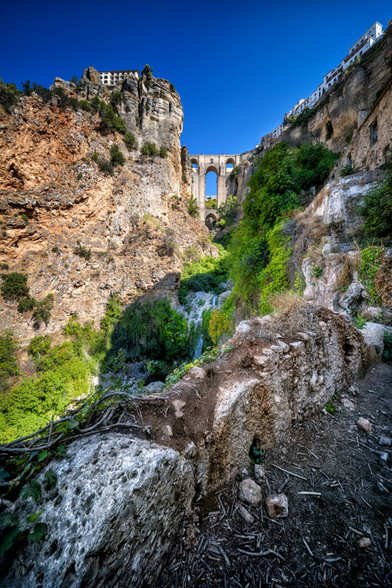 Ein tief eingeschnittener Felsencanyon mit üppigem Grün an den Hängen. In der Mitte führt eine hohe, steinerne Bogenbrücke über die Schlucht. Links und rechts ragen steile Felswände empor, an denen sich Häuser und Ruinen anschmiegen. Im Vordergrund fliesst ein kleines Rinnsal über moosbewachsene Steine und Baumwurzeln. Der Himmel darüber ist klar und blau.
