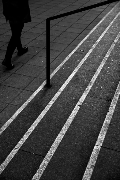 Oblique view of a long staircase with four steps in an urban setting. The edges stand out in stark contrast to the dark flooring with a white stripe. In the upper left half, you can see a businessman in a dark suit walking by; more precisely, you can see his torso.