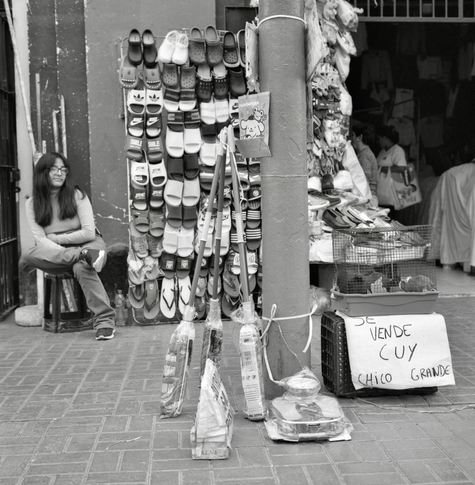 A black-and-white film photo. A woman is seated outdoors, selling shoes and other things in Lima, Peru. Thursday, September 18, 2025.