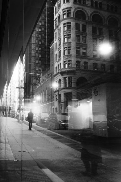 Two silhouettes are seen through a reflection of a window. Behind the silhouettes is the street, cars, a large stone corner building. Below them is the sidewalk. This is all blended with the interior of the window: a bare empty retail space.