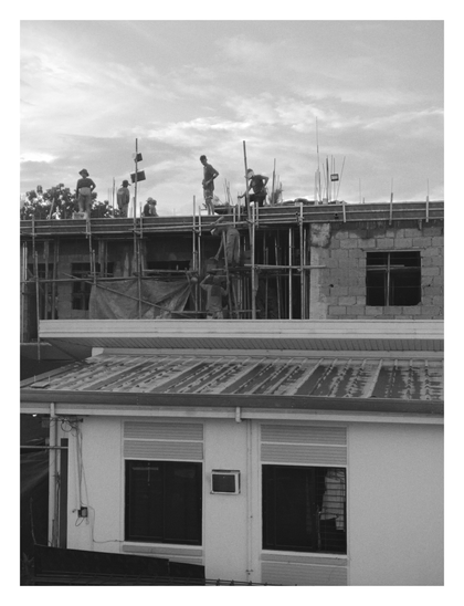 In the upper half, several workers are active on the top level of a building under construction, silhouetted against a cloudy sky. The structure features exposed block work, rebar, and scaffolding. The lower half of the image shows the corrugated metal roof and white facade of a finished building in the foreground. - Google Gemini 2.5 Pro