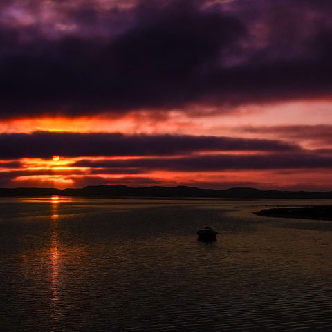 The image depicts a body of water, likely a lake or bay, at sunset. The sky is filled with layers of orange and red clouds, creating a dramatic and vibrant effect. The water reflects the colors of the sky, appearing golden and shimmering. A silhouetted landmass is visible in the distance, and a small dark shape, possibly a boat or a structure, is centered in the water near the foreground. The overall scene is dark with a strong emphasis on the warm colors of the sunset and the reflections on the water surface.