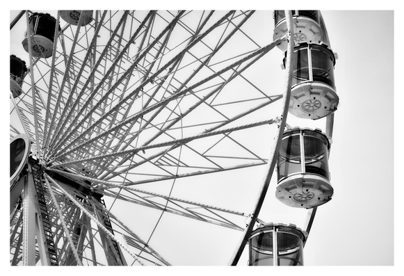 This is a black and white photograph capturing a large Ferris wheel from a low angle, looking upward toward the sky. The composition focuses on the wheel's intricate metal framework, with numerous spokes radiating outward from the central hub like the rays of a sun. On the right side, several enclosed passenger cabins—cylindrical and glass-paneled—are visible, stacked along the curving rim, giving a sense of height and motion. The background is a plain, overcast sky, which emphasizes the stark, geometric lines of the structure and creates a dramatic, almost industrial atmosphere. The image evokes a feeling of scale and engineering wonder, with no people or additional elements present.