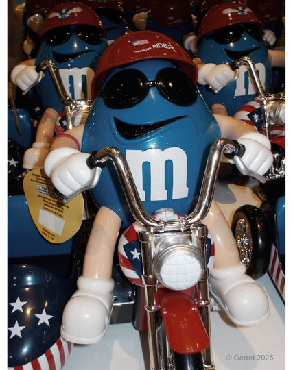 Blue M&M character wearing sunglasses and a red "Freedom Rider" helmet, sitting on a chrome motorcycle with stars and stripes, exuding a cool, fun vibe.