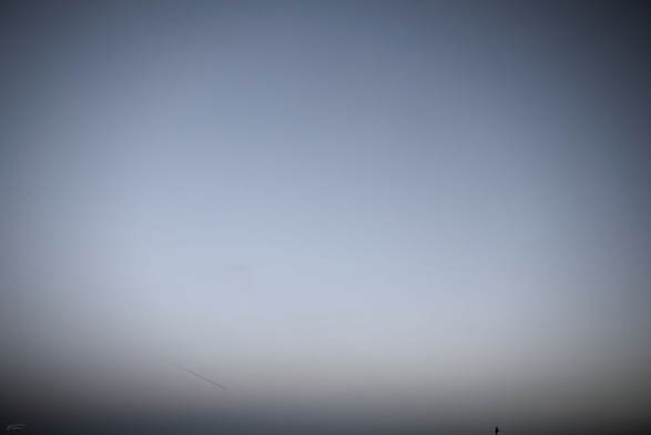 Das Foto zeigt eine extrem minimalistische und ruhige Szene: Ein fast vollständig in einem sanften Grau- und Blauverlauf gehüllter Himmel dominiert das Bild. Der obere Bereich ist heller und bläulich, während der untere Bereich dunkler und grauer wird. Im unteren Drittel des Bildes ist ein dünner, horizontaler Streifen zu sehen, ein Kondensstreifen eines Flugzeugs. Am unteren Rand des Bildes ist eine kleine, dunkle Silhouette mit zwei roten Lichtern zu erkennen. Es sind nur die obersten Meter eines hohen Industrieschornsteins. Das Bild wirkt sehr abstrakt und fast meditativ.