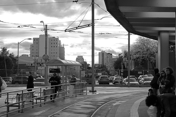 Late afternoon, after rain. In the upper right is the overhang of the roof of the outdoor platform. Lower right, several riders wait for the arrival of their streetcar. Lower left is the waiting area for another streetcar line with its own riders around a small glass shelter. In the upper left is a moody sky with the remnants of rainclouds over the tall buildings in the distance. 