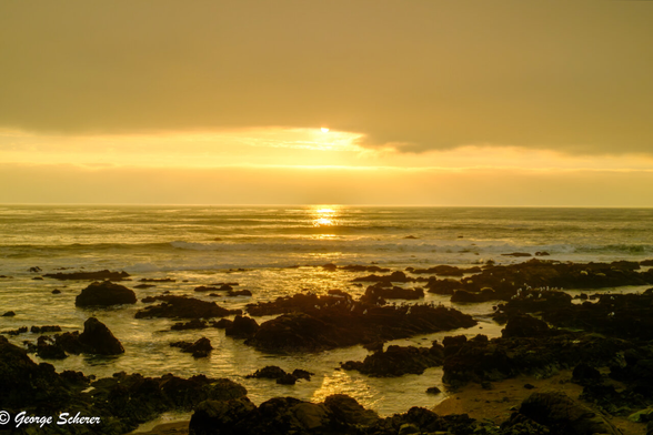 As it descends at the end of the day, the sun is emerging from low clouds, illuminating the waves on the ocean.  There is a rocky beach in the foreground.  The sun's illumination gives everything in the scene a golden glow.