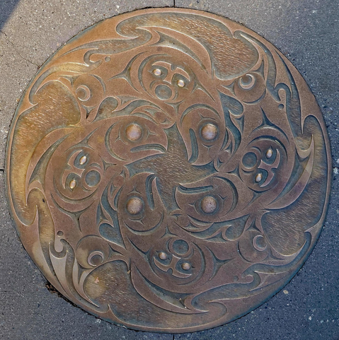 Coast Salish storm drain design by Musqueam artist Susan Point. The bronze cover depicts salmon in a circular cycle with human beings. Brighter portions of bronze have been polished by pedestrians. 